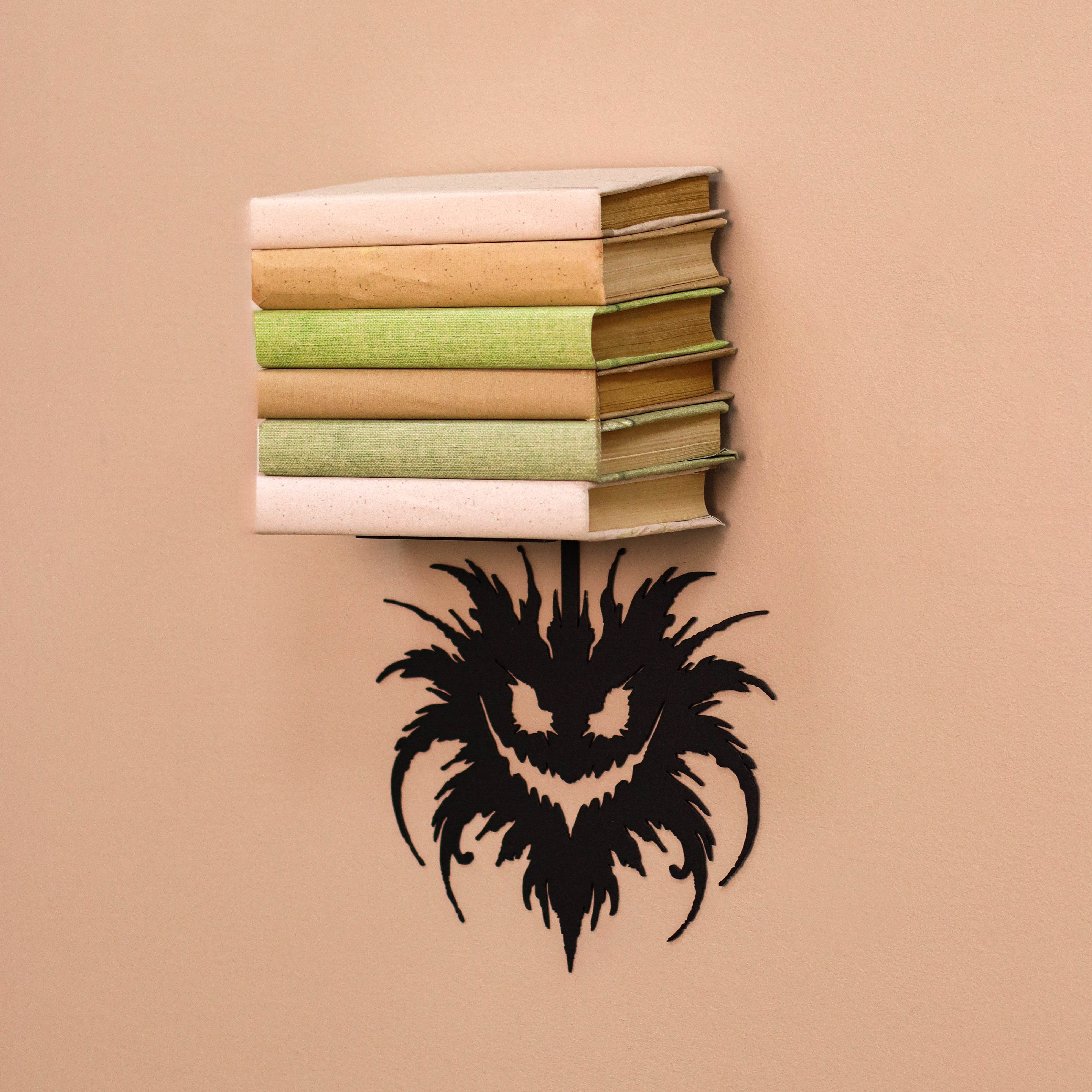 A stack of five decorative books with beige, green, and cream spines is mounted on a warm beige wall above a black, stylized monster face cutout.