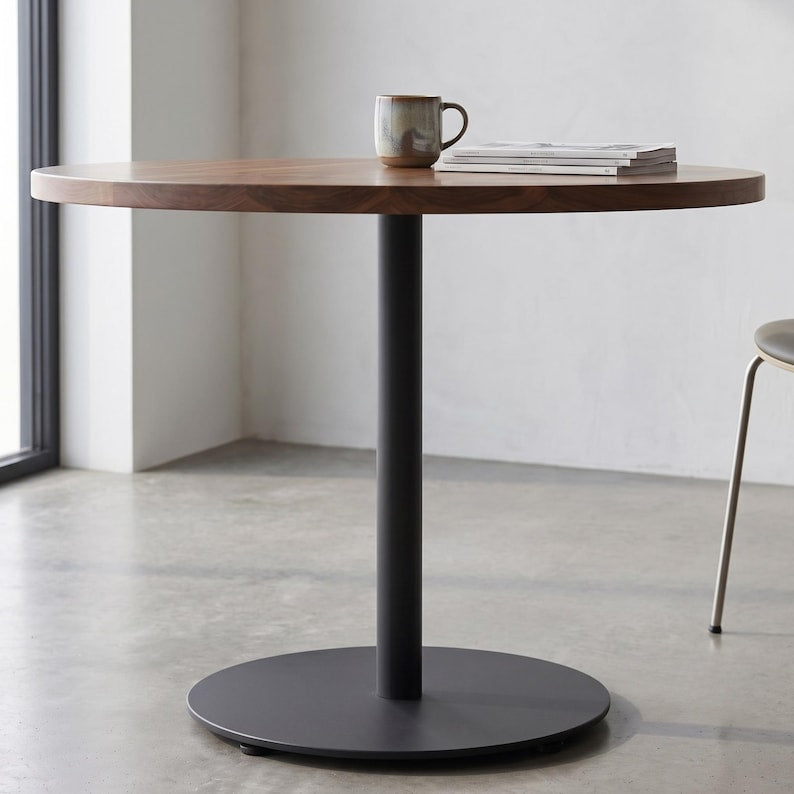 A round wooden dining table with a black metal pedestal base and circular footplate, styled with a ceramic mug and magazines on a light gray concrete floor in a minimalist room.