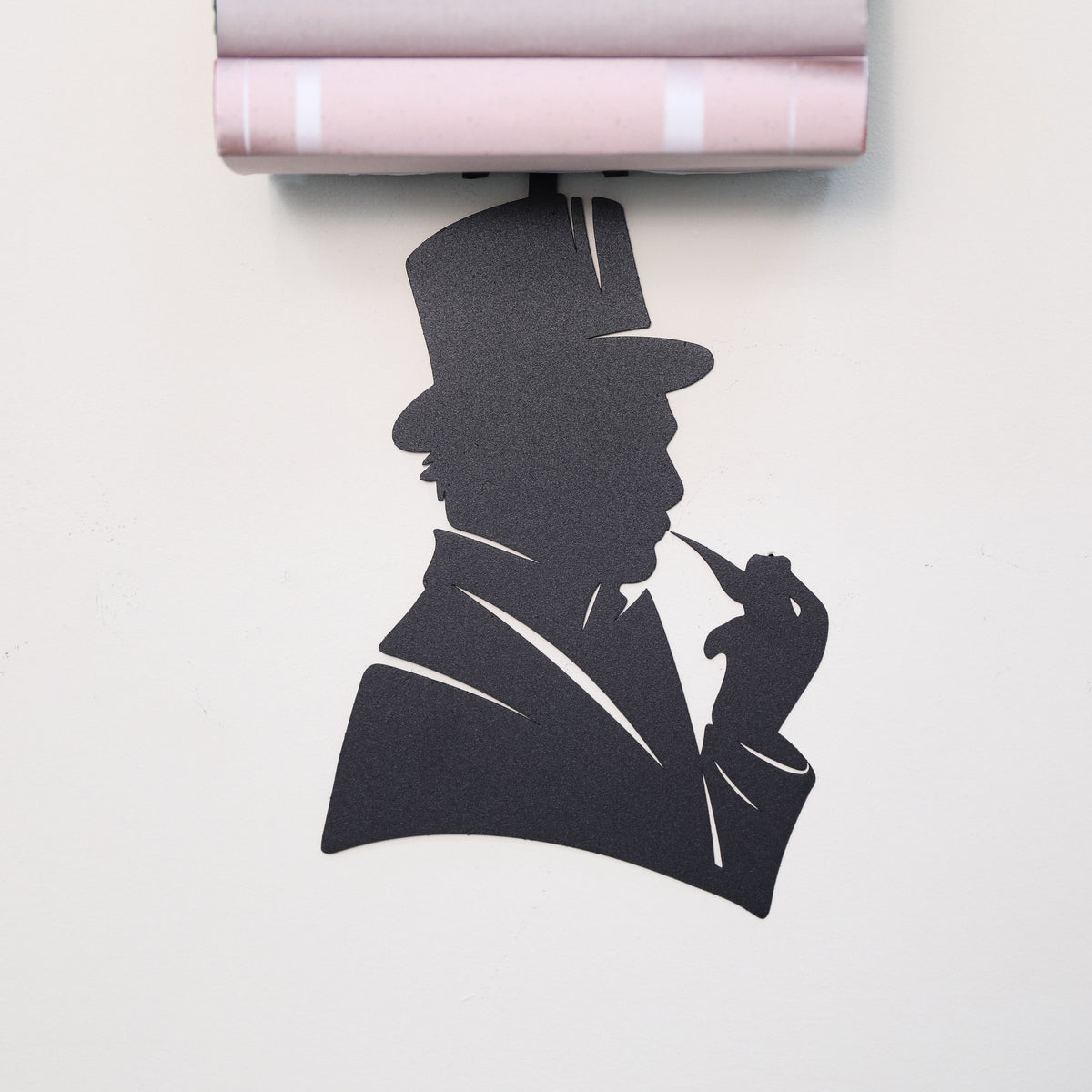 A black silhouette wall art piece shaped like a man in a top hat and suit holding a pipe, shown against a light wall with a rolled sheet of pink patterned paper mounted above it.