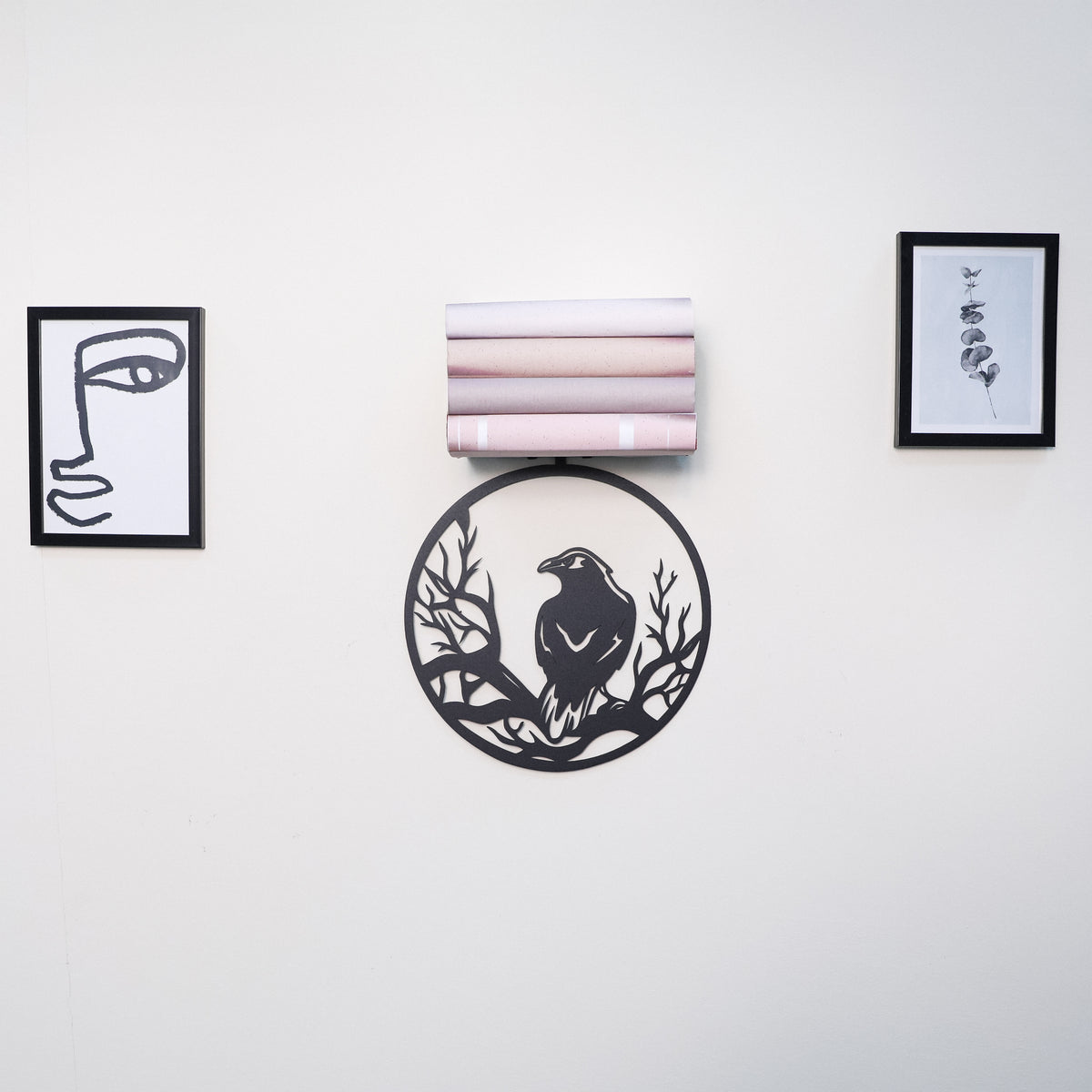 A wall-mounted decorative shelf or organizer with five horizontal tiers holding folded light pink and white textiles, centered on a white wall. Below the shelf is a circular black metal wall art piece featuring a bird silhouette.