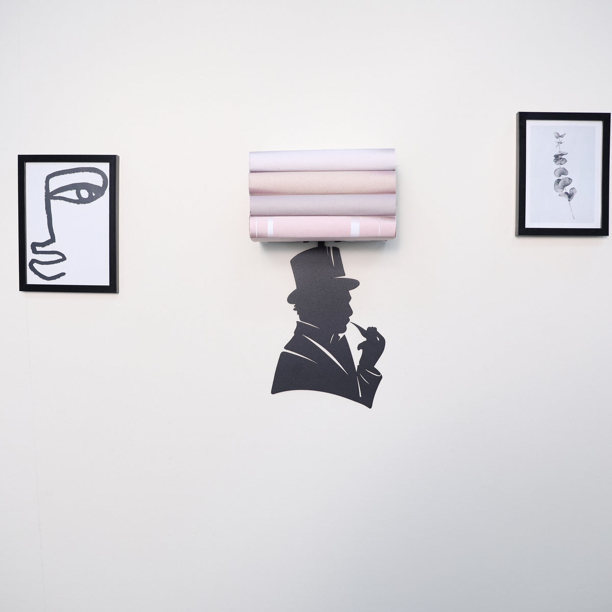 A wall-mounted shelf holding five rolled sheets of paper in soft blush, pink, and beige tones, positioned centrally between two black-framed abstract art prints on a white wall.
