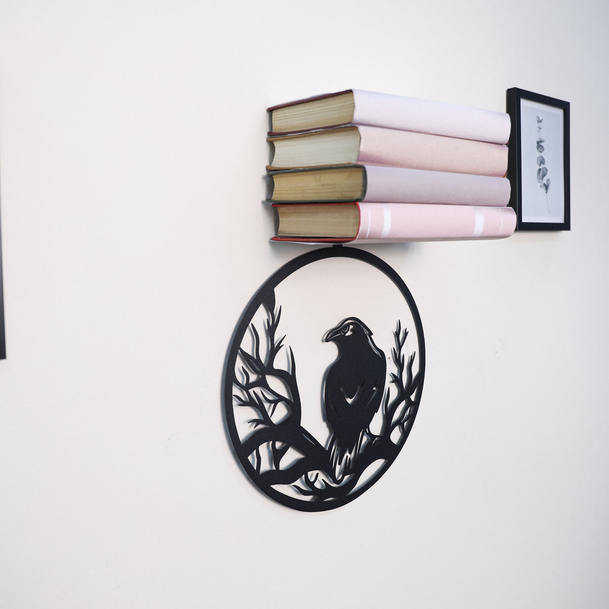 A black metal wall decor piece shaped like a raven perched on branches, mounted on a white wall below a floating bookshelf holding pink and beige books and a small framed print.