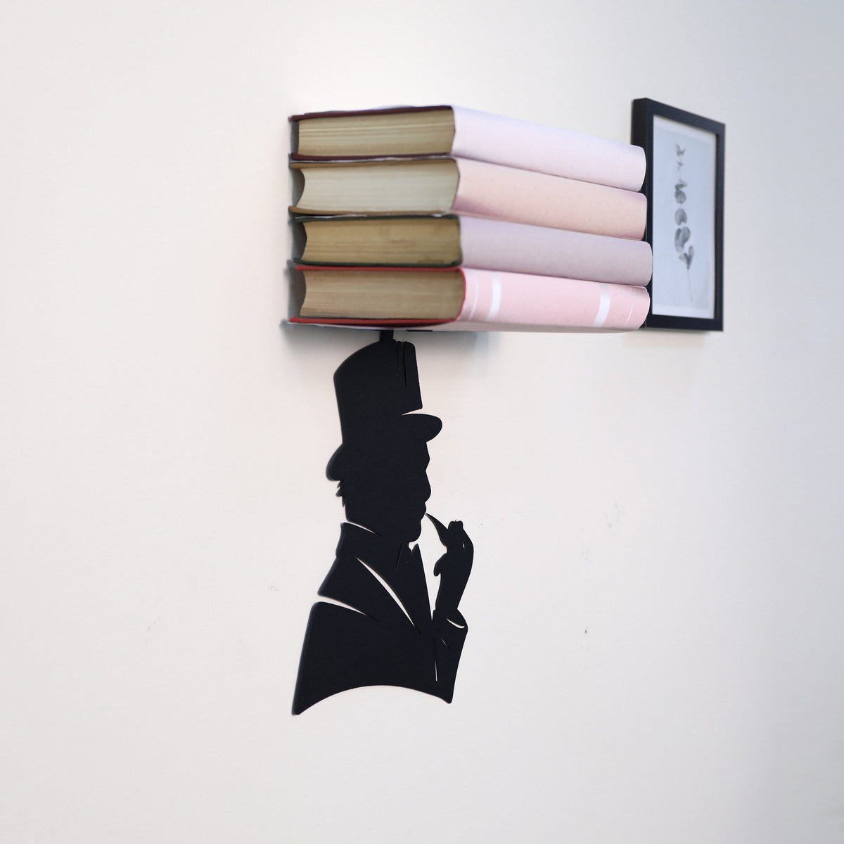 A wall-mounted bookshelf shaped like a stack of books with a small framed print, shown against a plain white wall. A black silhouette of a man in a top hat and suit appears below the shelf.