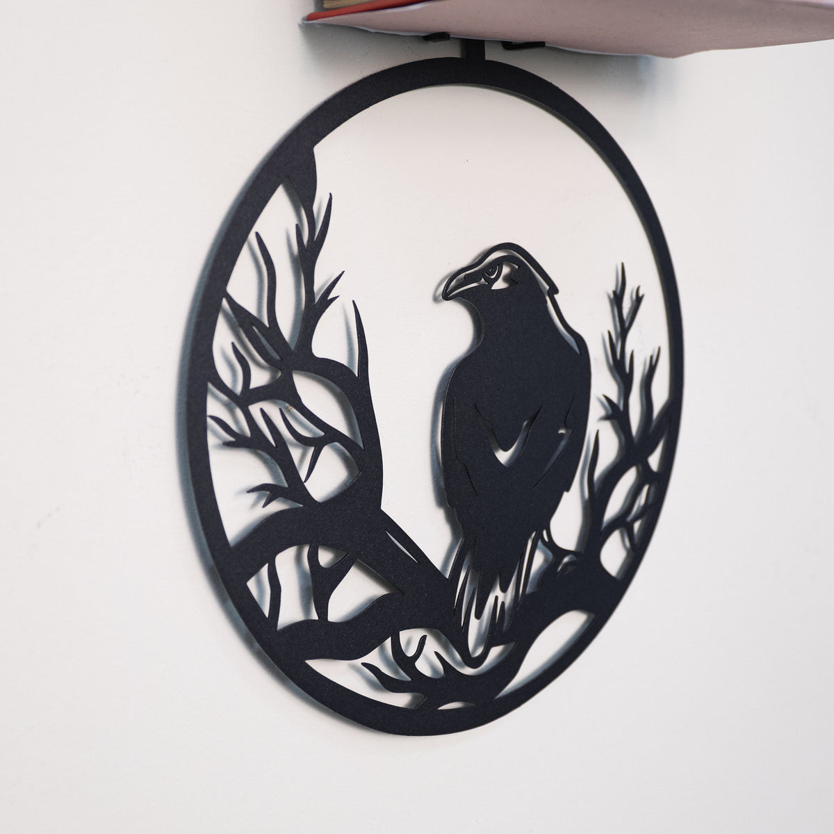 A black metal wall art piece featuring a raven perched among bare tree branches inside an oval frame, mounted on a plain white wall.