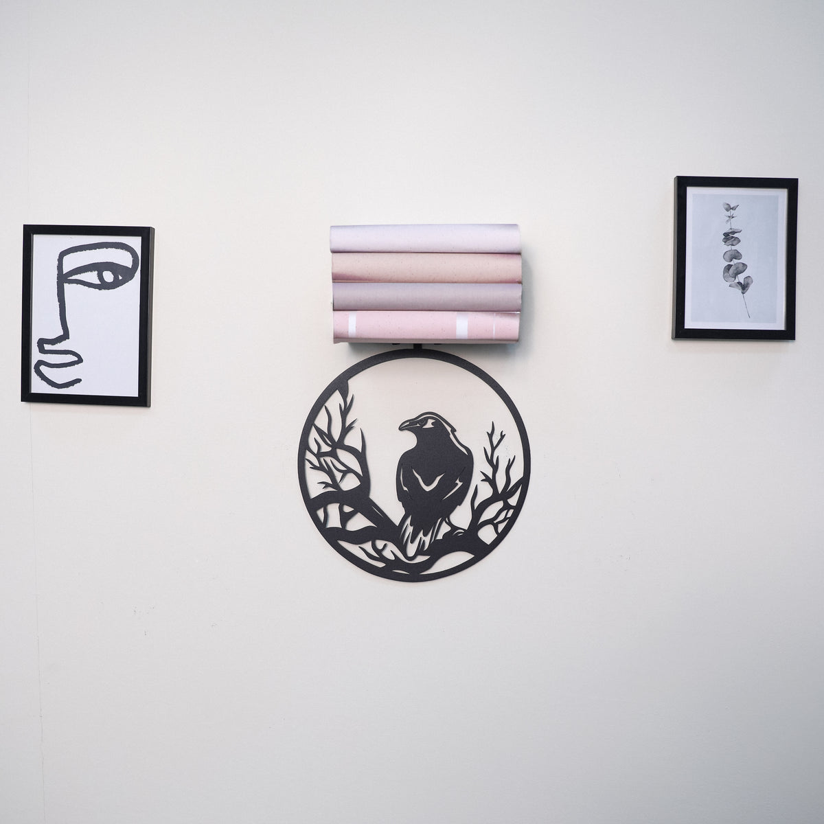 A wall-mounted decorative arrangement on a light neutral wall, featuring a black circular cutout with a bird and branch motif, a stack of pastel rolled sheets, and two small framed abstract prints.