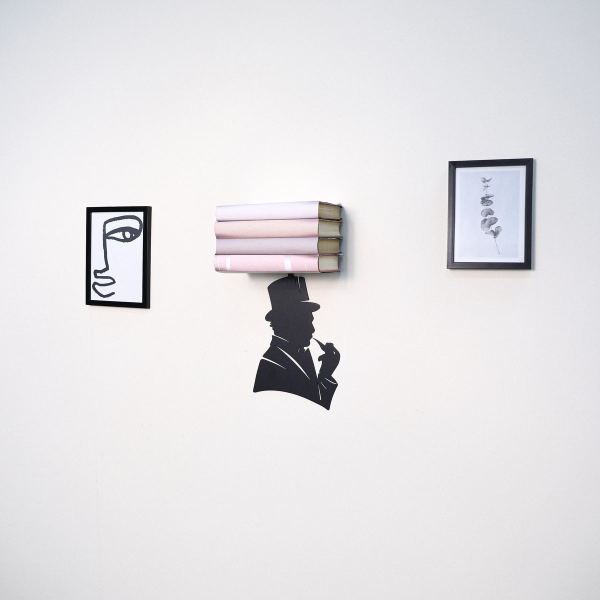 A wall-mounted bookshelf shaped like a stack of pink and beige books, with a black silhouette of a man in a top hat hanging from the center. Two small framed black-and-white art prints hang on either side against a plain white wall.