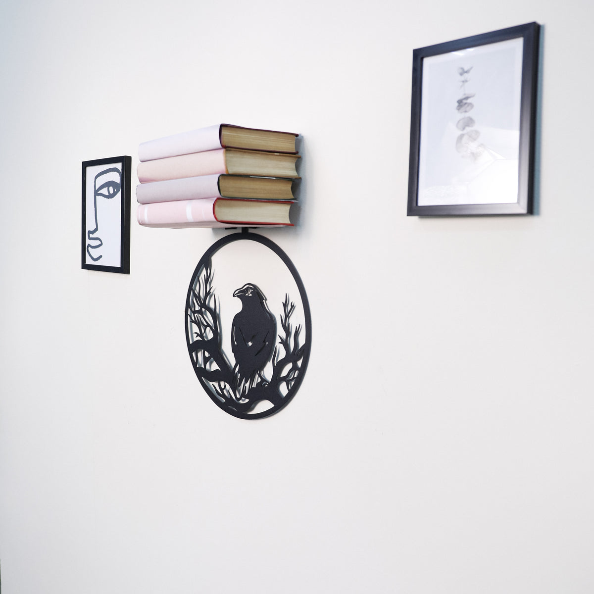 A black metal wall-mounted bookshelf shaped like an oval with a raven silhouette, holding a stack of books, shown on a white wall alongside two framed art prints.