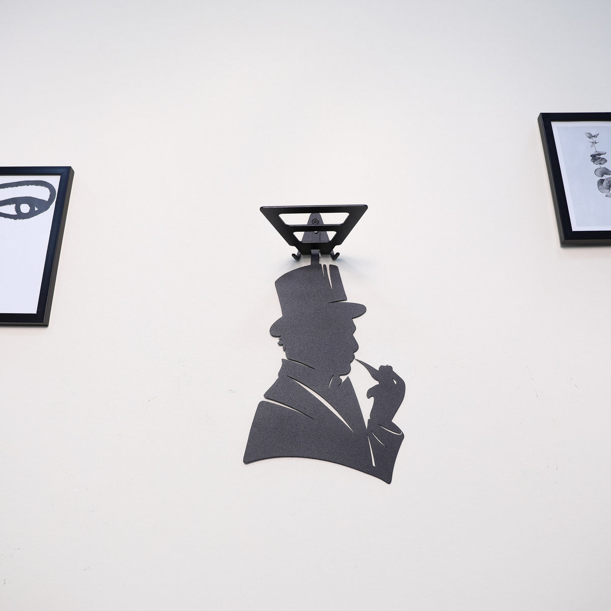 A black metal wall-mounted silhouette shelf featuring a stylized figure in a top hat and suit holding a cigarette, mounted on a white wall with framed art partially visible on either side.
