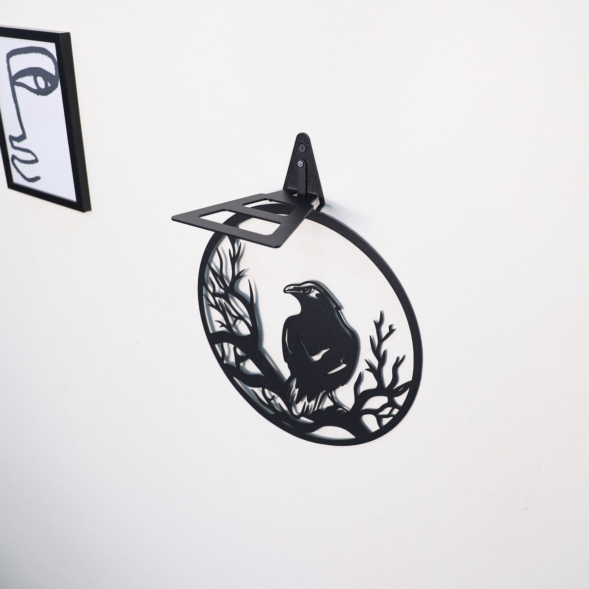 Black metal wall-mounted shelf with a circular cutout design featuring a raven perched on branches, shown against a plain white wall.
