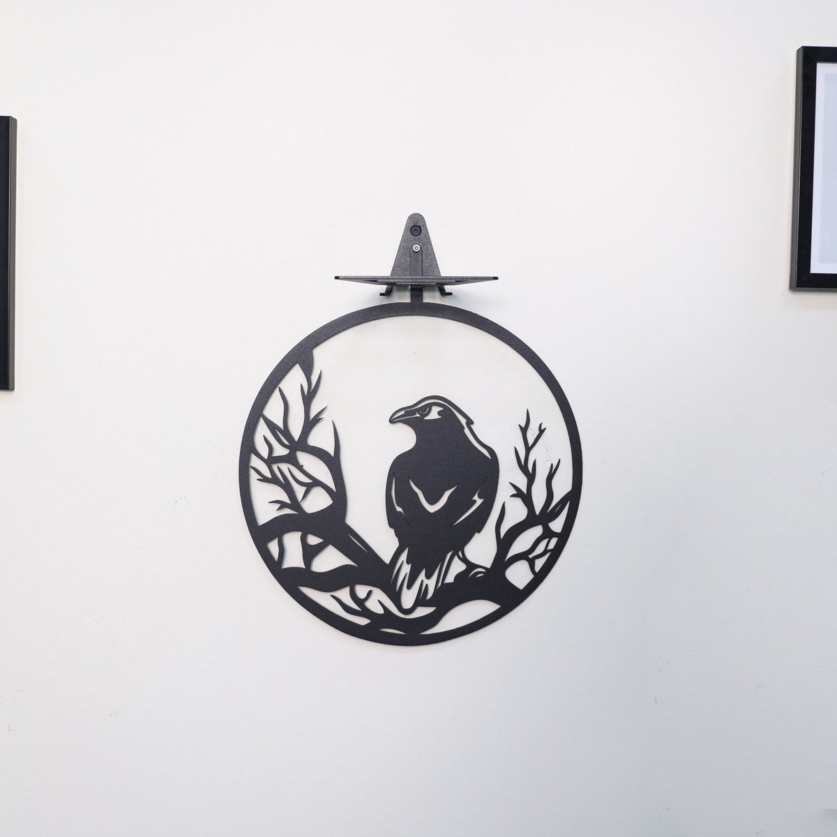 A black metal wall art piece featuring a raven perched on branches inside a circular frame, mounted on a white wall.
