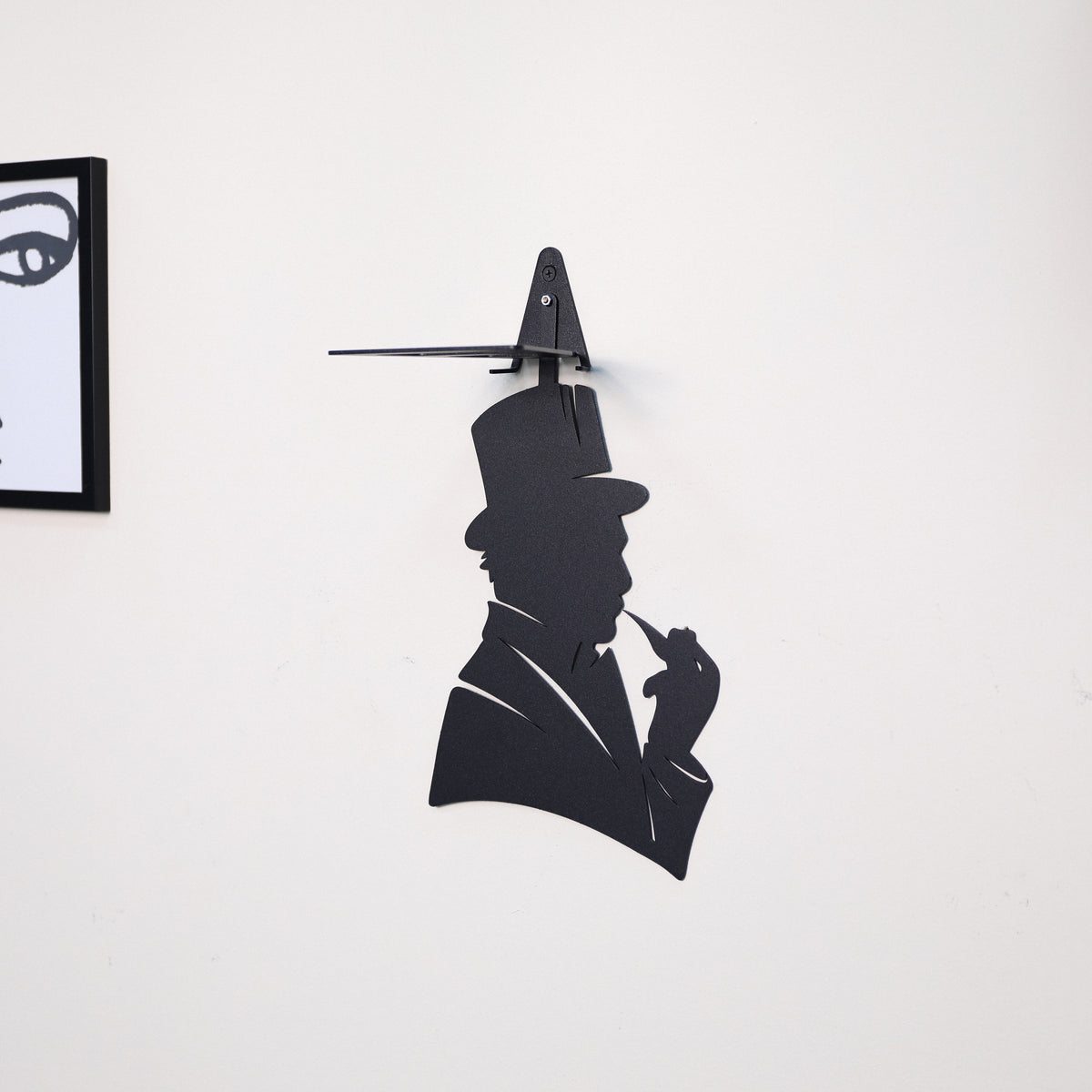 A black metal wall art piece shaped like a man in a top hat smoking a pipe, mounted on a plain white wall. Part of a framed black-and-white print is visible at the left edge.