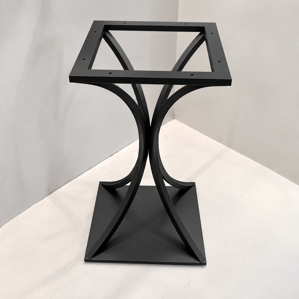 A black metal side table base with a square top and curved intersecting legs, photographed against a plain light gray corner backdrop.