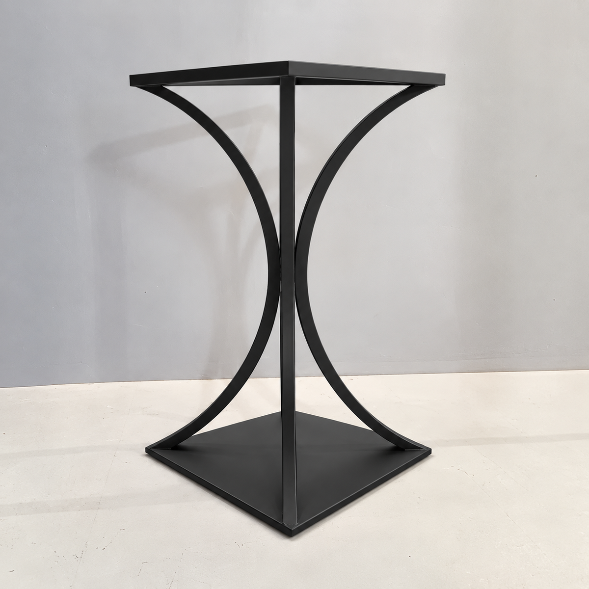 A modern black metal side table featuring a square top, a central vertical post, and two sweeping curved legs that form an hourglass shape over a square base, set against a plain light gray wall and white floor.