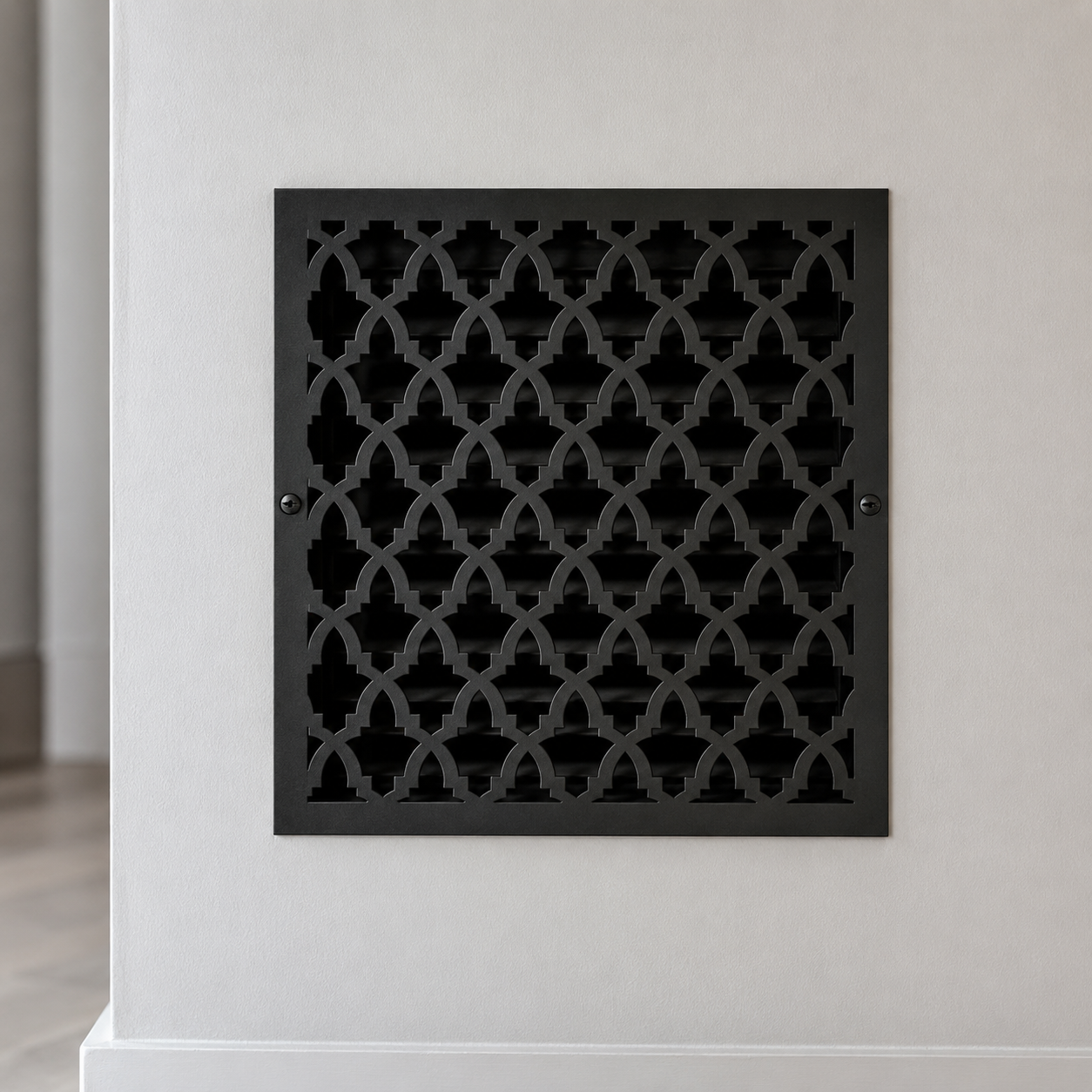 A square black metal vent cover with an ornate repeating trellis pattern, mounted on a light gray wall.