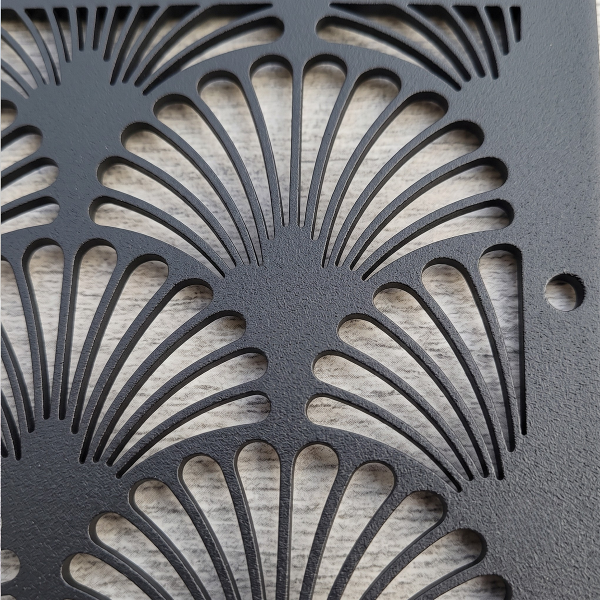 A close-up of a black metal decorative panel with a repeating fan-shaped cutout pattern, shown over a light wood surface.